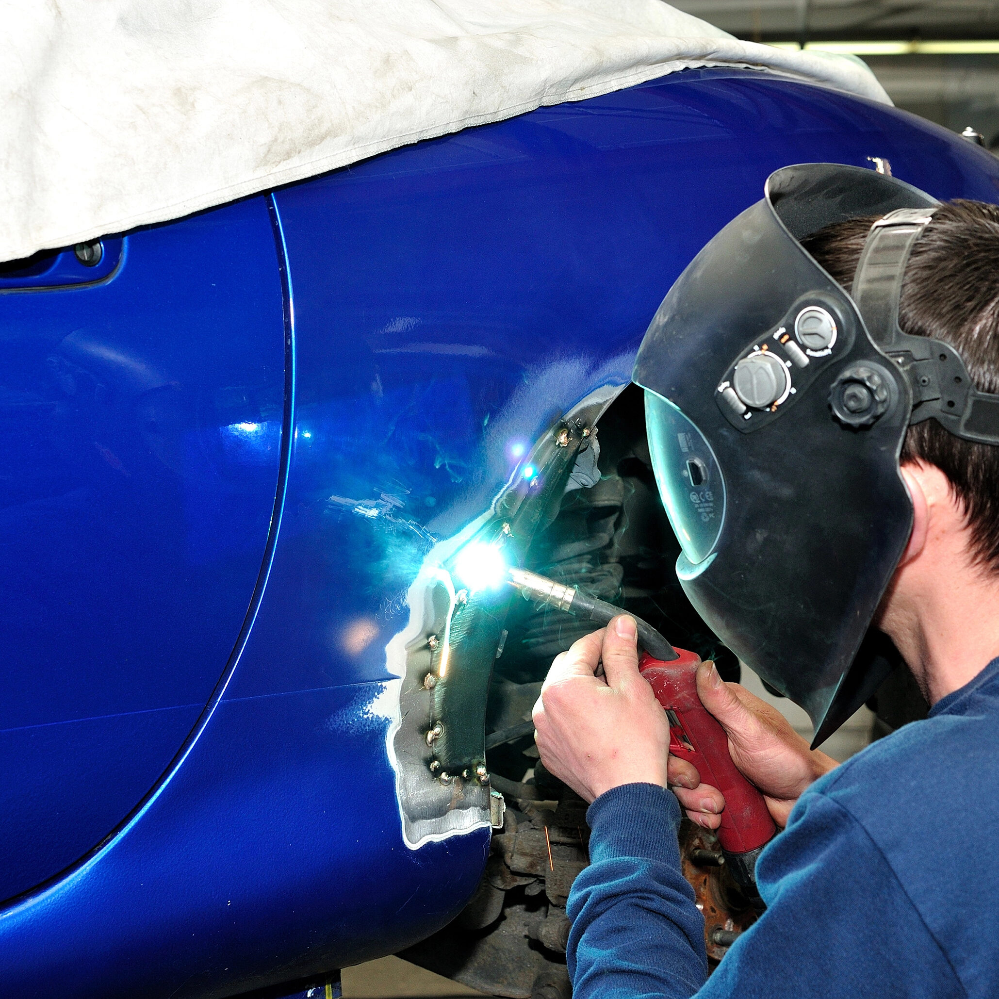 A mobile welder, donning a welding helmet, is expertly welding a patch on the side of a blue car in the workshop. The welding torch emits a bright light, illuminating the bare metal that contrasts sharply with the painted surface.
