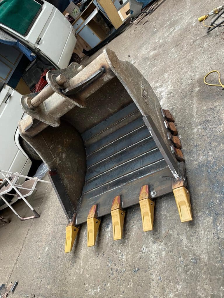 An excavator bucket with yellow teeth attachments placed on a workshop floor. A white van and various tools are in the background, along with a chair and coiled extension cord.