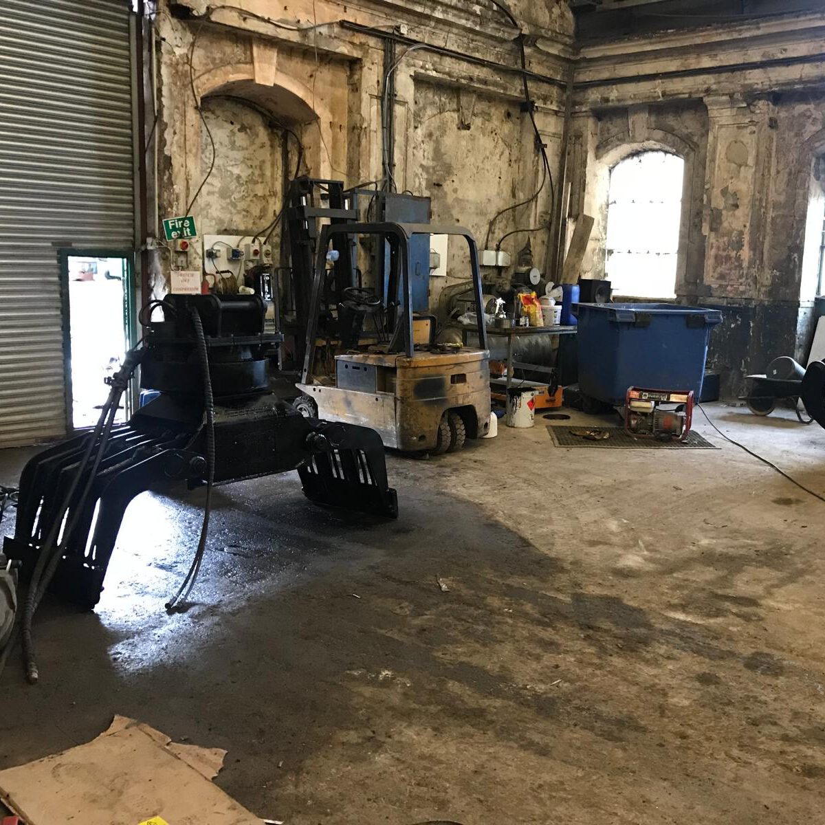 A dimly lit, worn-out industrial workshop with a forklift, a large blue container, and two heavy-duty metal claw attachments on the ground. The walls are aged, and there's a partially open roller shutter door on the left.
