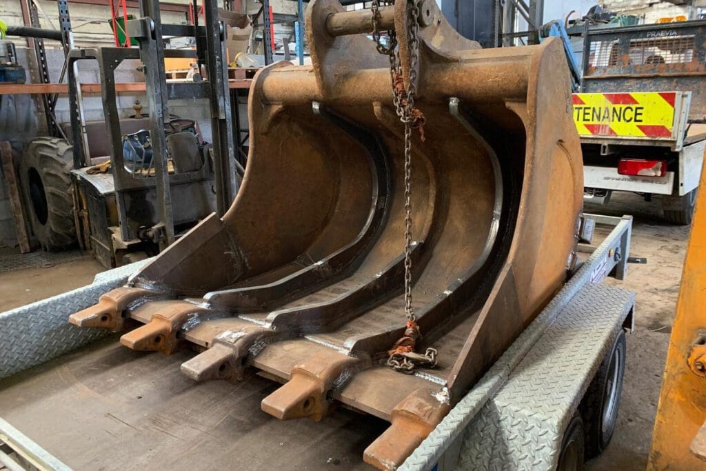 A large, rust-colored excavator bucket with three sections is secured to a trailer with chains. The background shows a cluttered workshop with tools, equipment, and a vehicle displaying a "Maintenance" sign.