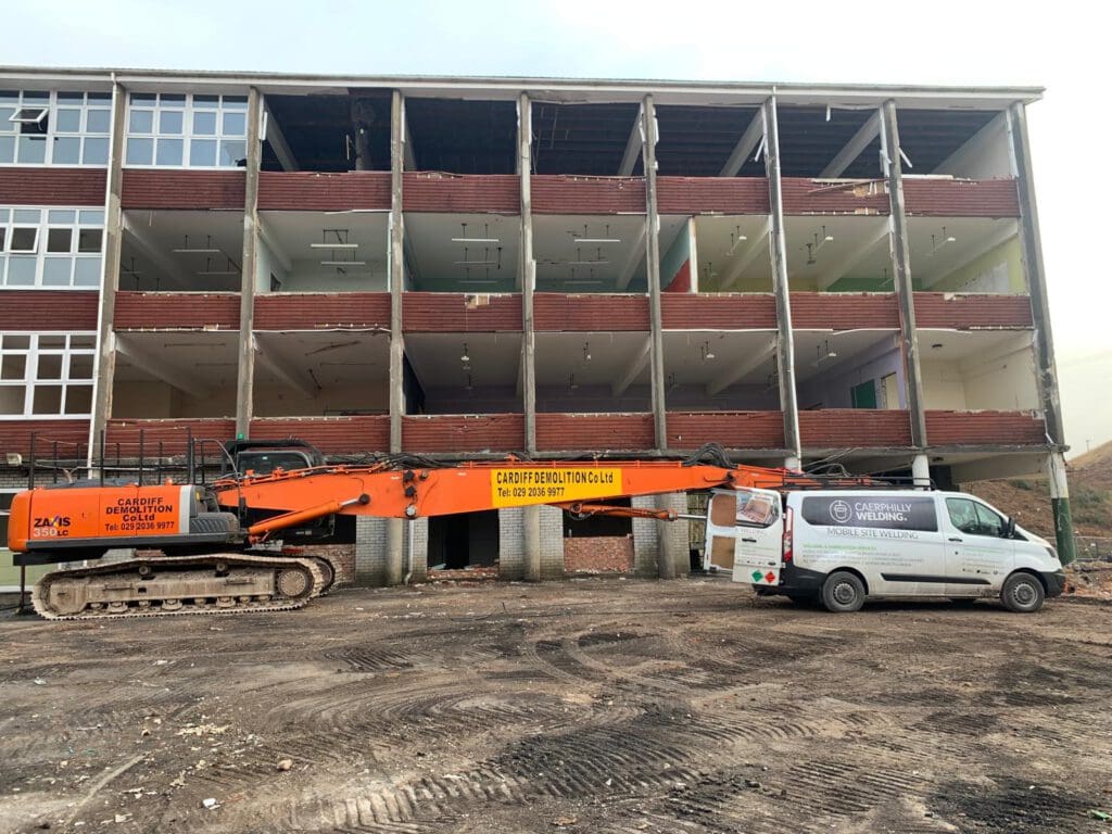 A partially demolished multi-story building with exposed floors and debris. A large orange excavator and a white van from Cardiff Demolition are parked in front. The ground is covered with dirt and rubble.