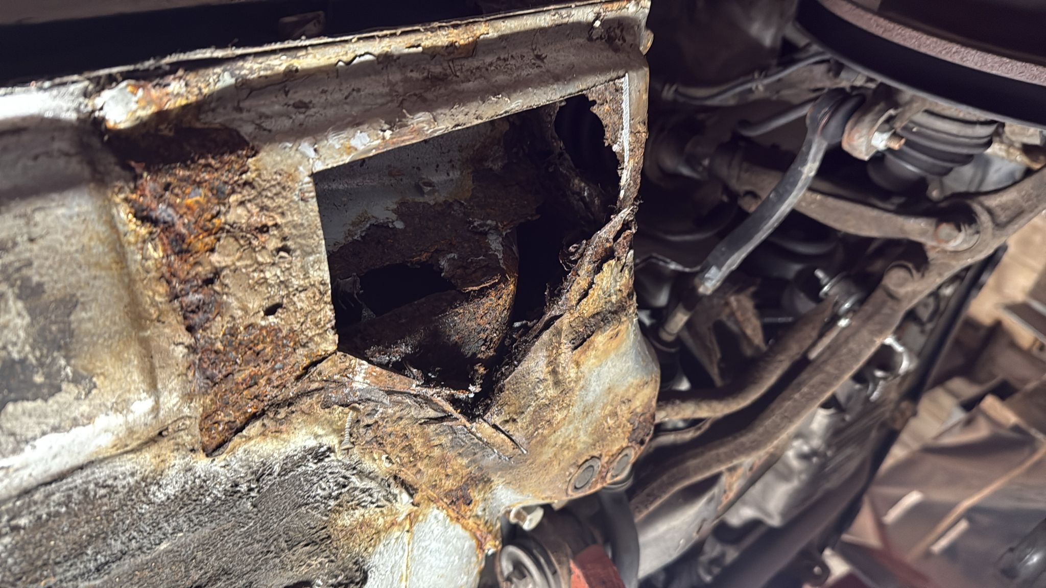 A close-up of a car chassis with severe rust damage and a large hole, exposing the corroded metal and interior structure near the engine and suspension components.