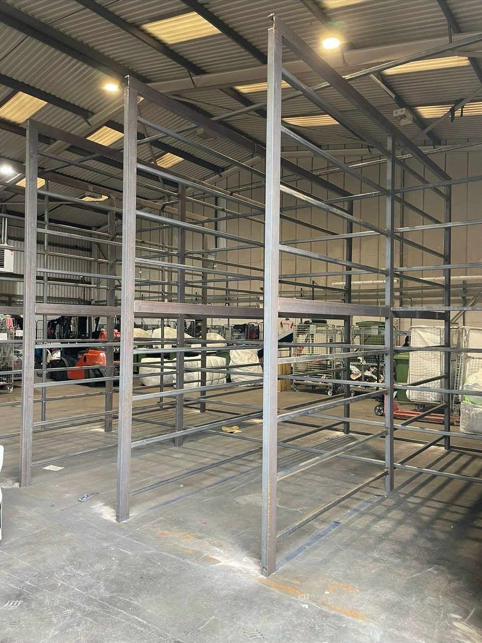 In a spacious warehouse with high ceilings, a large metal industrial shelving unit is taking shape. Mobile welders and plant repair equipment are visible in the background, highlighting the unfinished metal beams. Bright lighting showcases the floor and modern construction environment.
